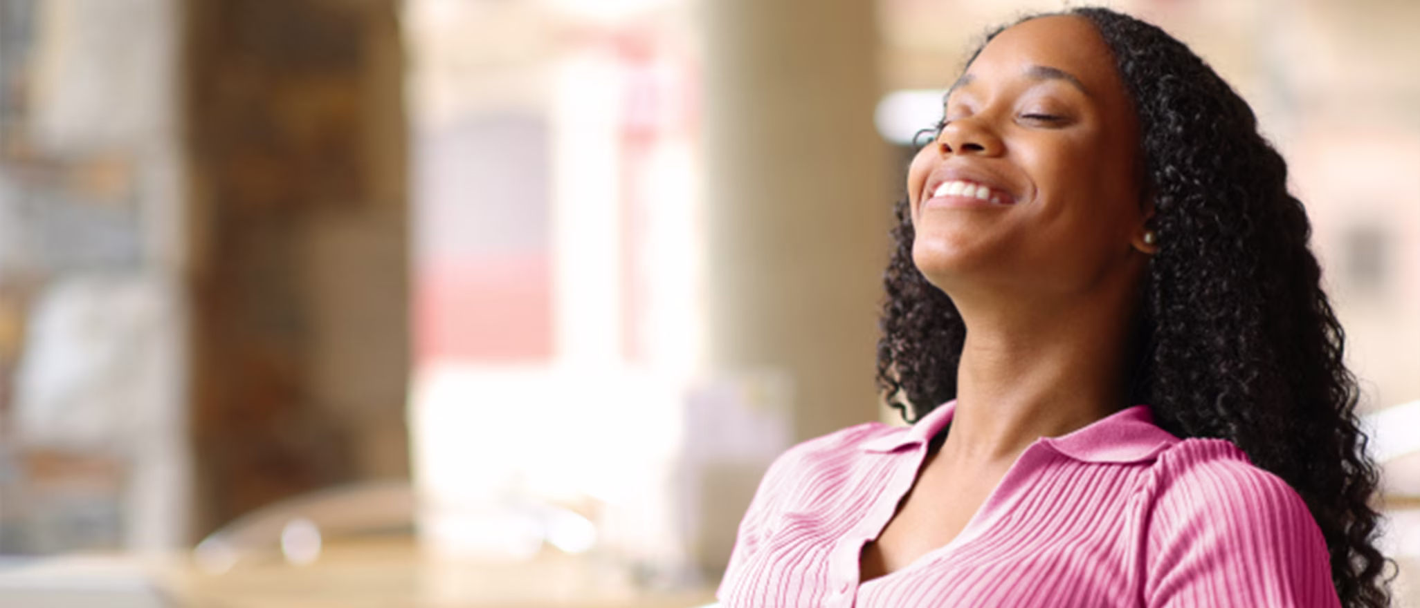 A woman sitting outdoors, closing her eyes and smiling with relief as she enjoys the sun.