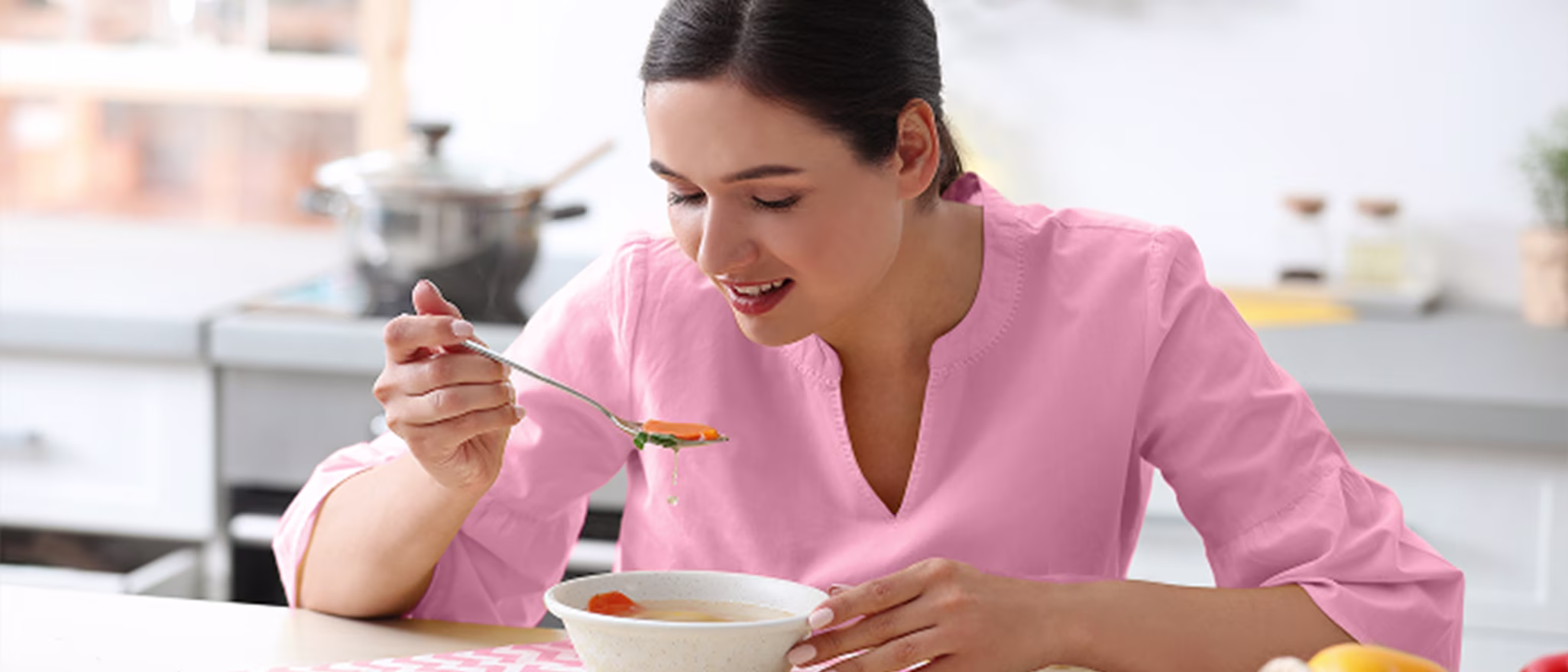 A woman in a pink shirt sitting at a kitchen table, happily eating a bowl of vegetable soup.