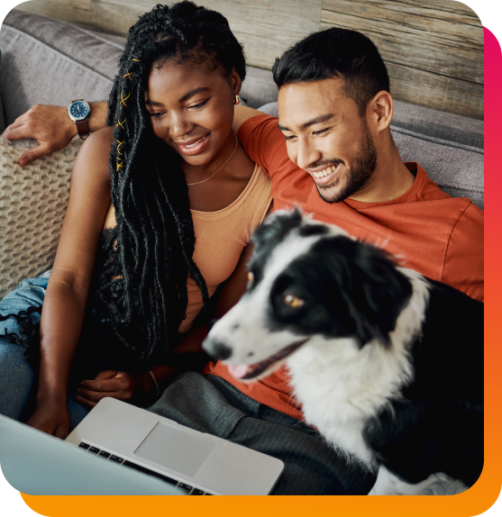 Smiling couple sitting on couch with dog looking at a laptop.