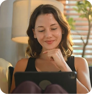 White woman with shoulder-length brown hair, wearing a black sleeveless top and smiling while using a tablet at home.