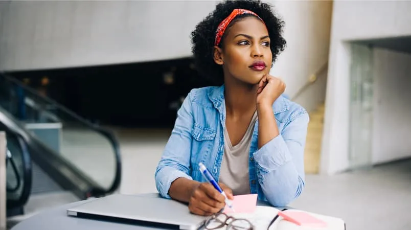 Woman with computer and notebook at table looking away with pen in hand