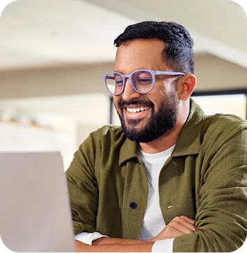 Man with short dark hair, a beard, and glasses, wearing an olive-green overshirt and smiling while using a laptop.