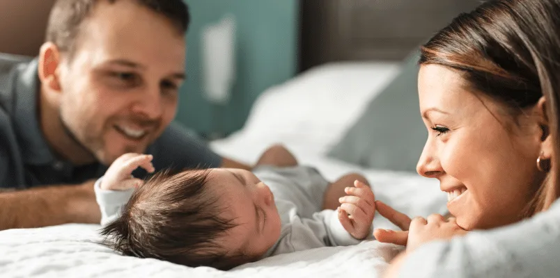 Parents smiling at their baby on the bed while they contemplate opening up a trump account for the child.