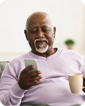 Older Black man with a gray-and-white goatee, wearing a light-purple sweater and holding a coffee mug while looking at his smartphone