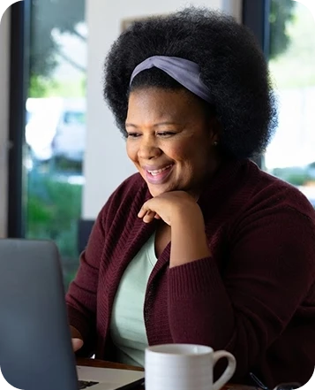 Black woman wearing a lavender headband and burgundy cardigan, smiling while using a laptop at a table.