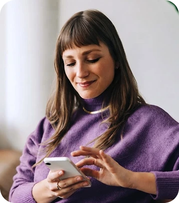 White woman with long brown hair and bangs, wearing a purple turtleneck sweater and smiling while using a smartphone.