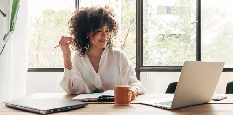 Woman at desk with two laptops a coffee cup and a notebook