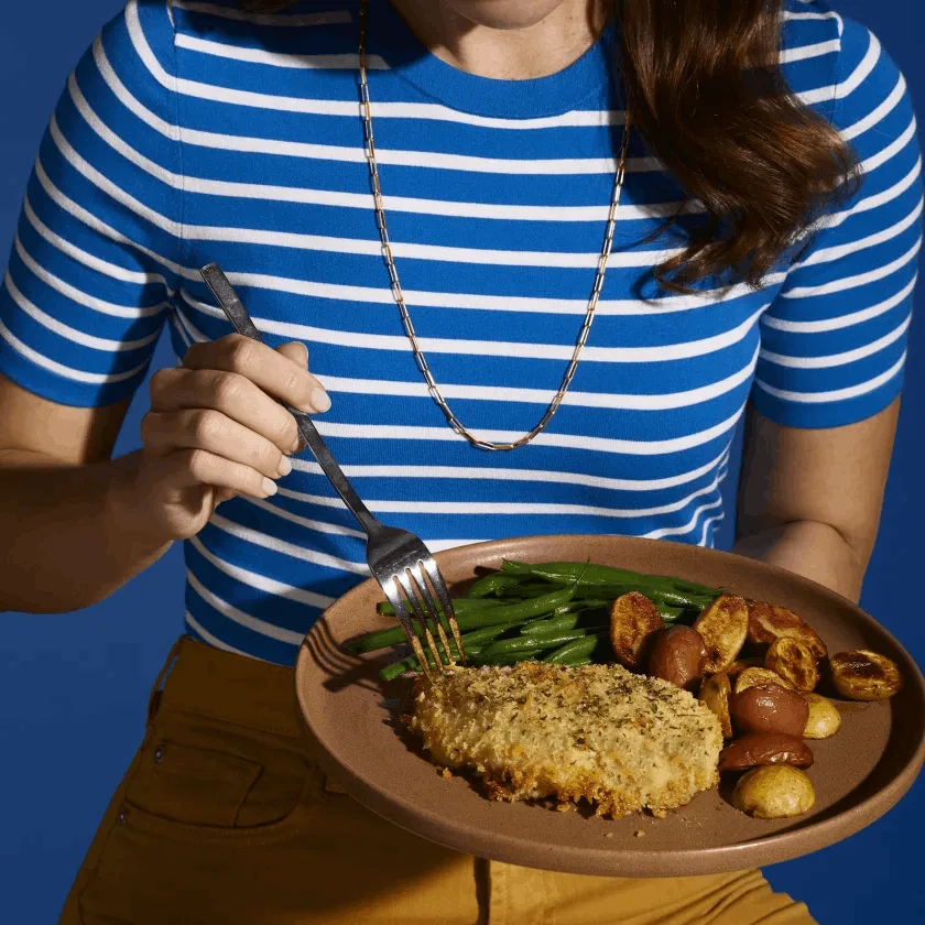 Woman in a striped blue shirt eating crispy chicken, roasted potatoes and asparagus on a brown plate