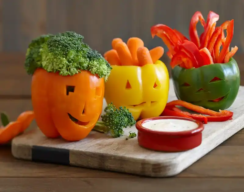 Orange, yellow and green bell peppers carved to look like Jack-o-lanterns on a wood cutting board with dipping vegetables and ranch.