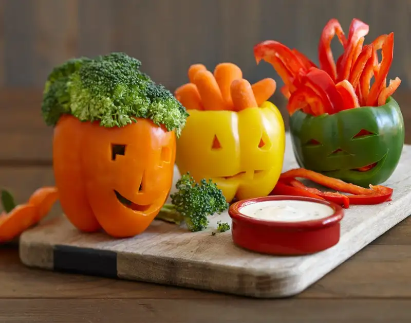 Orange, yellow and green bell peppers carved to look like Jack-o-lanterns on a wood cutting board with dipping vegetables and ranch.