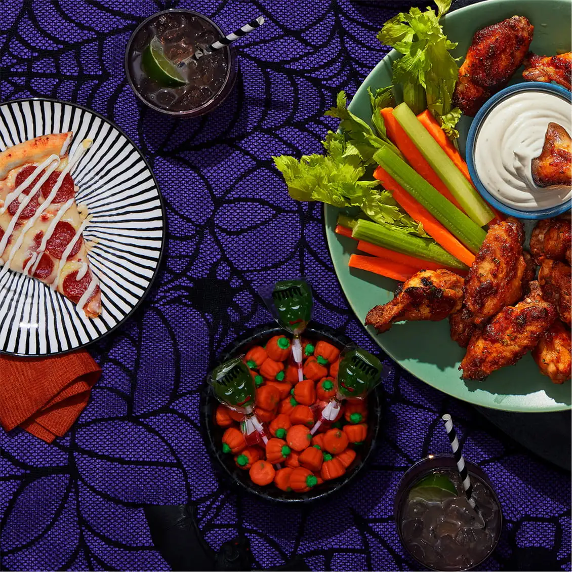 Halloween food layout with chicken wings, pizza slice, candy, and drinks on a purple spiderweb cloth