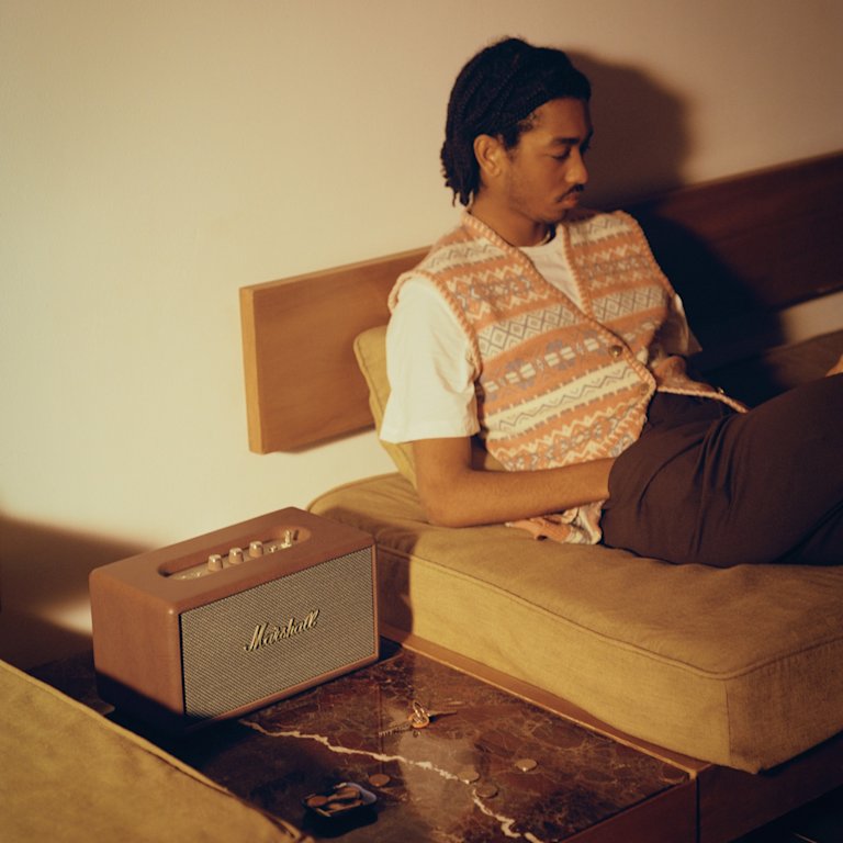 A man sitting on a couch next to a brown speaker, the Marshall Stanmore III Brown.