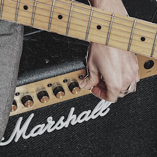 Close-up of a person's hand on an electric guitar neck, with a Marshall amplifier and control knobs visible in the background.