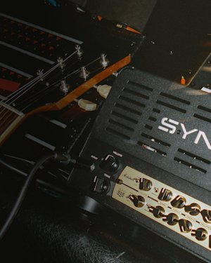 Nahaufnahme eines E-Gitarrenhalses neben einem Synergy-Gitarrenverstärkerkopf mit sichtbaren Reglern und Eingangsbuchsen.