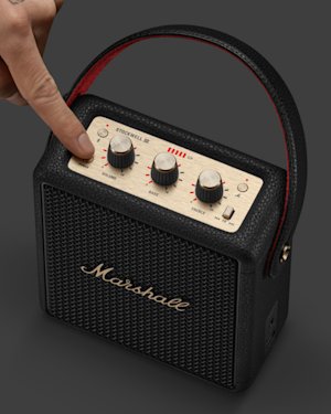 A hand pressing the volume knob on a black Marshall Stockwell II portable speaker with gold control panel and red-lined handle.