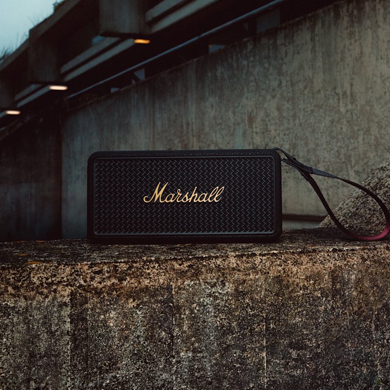 Marshall Middleton II Black and Brass portable Bluetooth speaker sits on a concrete ledge with a concrete structure and lights in the background.