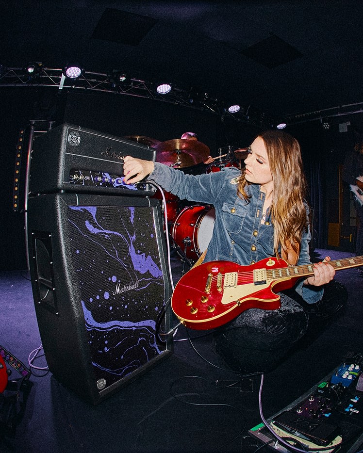 Woman adjusting the knobs on a Marshall Studio JMH Half Stack Hendrix Edition while holding a red electric guitar on stage, with drums and stage lights in the background.