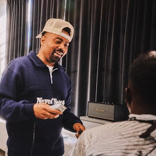 A man in a backwards cap and "Never Settle" sweatshirt holds a hair clipper; a Marshall speaker sits on a table nearby.