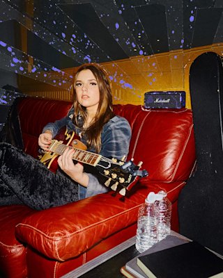 A woman sits on a red couch playing an electric guitar, with Emberton III Hendrix Edition speaker, a guitar case, notebooks, and water bottles nearby.