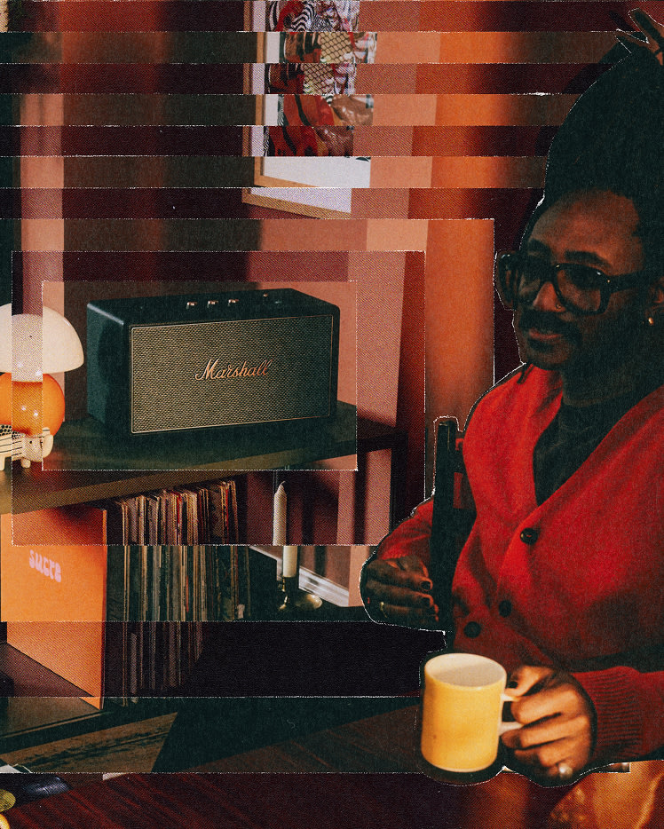 A person in glasses and a red outfit sits at a table in a room with a Marshall Stanmore IV Black speaker, vinyl records, and warm-toned decor.