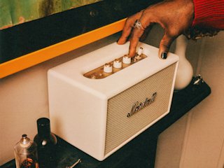 A person adjusts the knob on Acton IV Cream speaker placed on a black shelf next to decorative items.