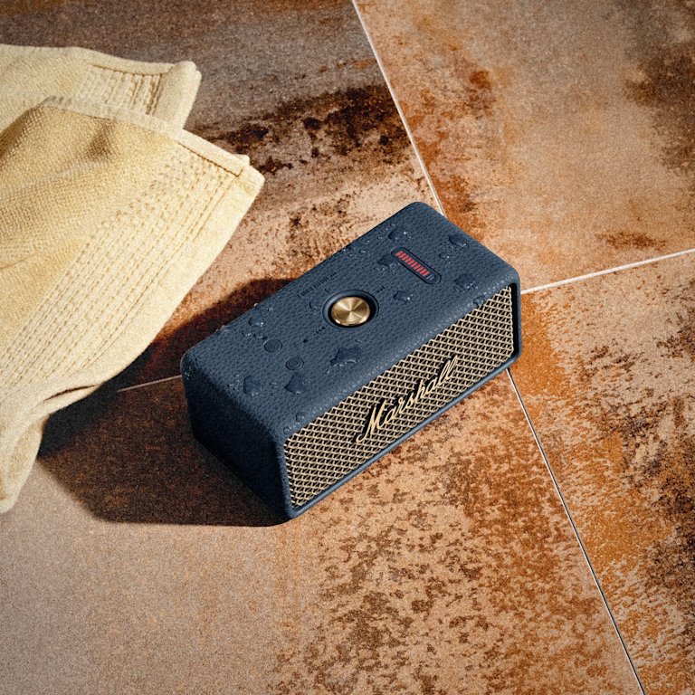 Marshall Emberton III Midnight Blue portable Bluetooth speaker sits on brown tiles next to a yellow towel, with water droplets visible on its surface.