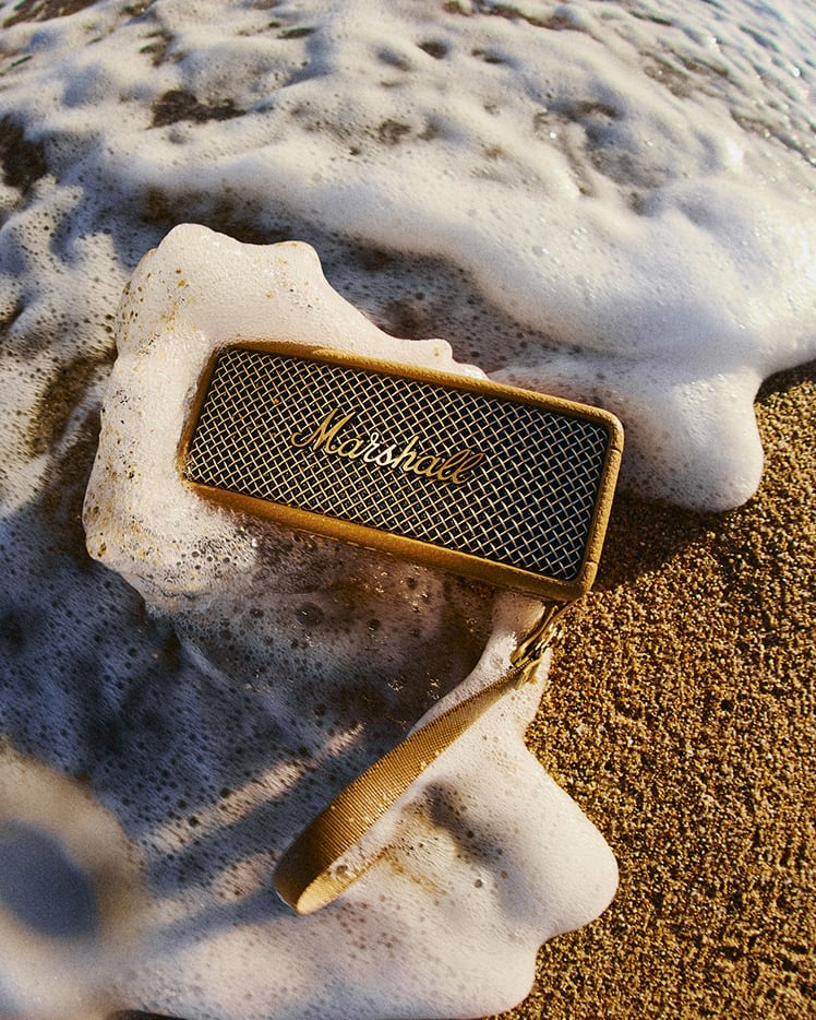 A Marshall Emberton III Brown portable speaker on wet sand is partially covered by ocean foam and water at the beach.