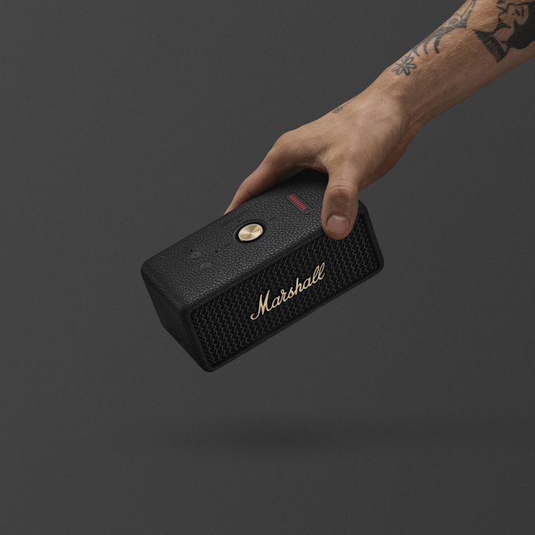 A hand holding Marshall Emberton III Black and Brass bluetooth speaker against a dark gray background.