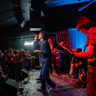 A band performs on stage in a dimly lit venue as an audience watches and photographs the show.