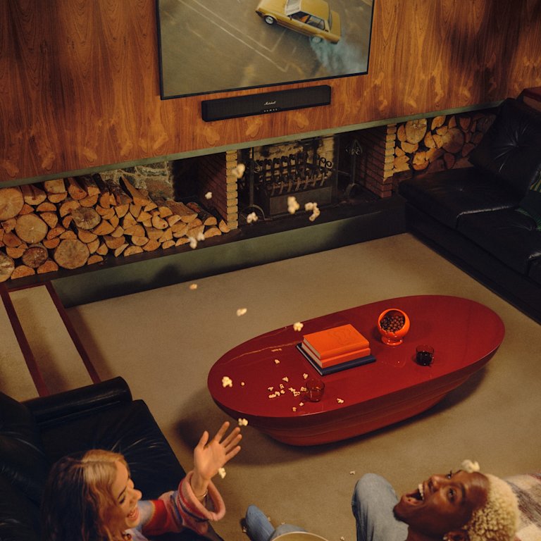 Two people relax on the sofa in a wood-paneled living room, watching a car TV with Marshall Heston 60 Black soundbar.