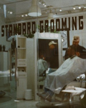 A barbershop interior is visible through a glass window with "Standard Grooming" written on it. A person sits in a chair while another stands nearby.