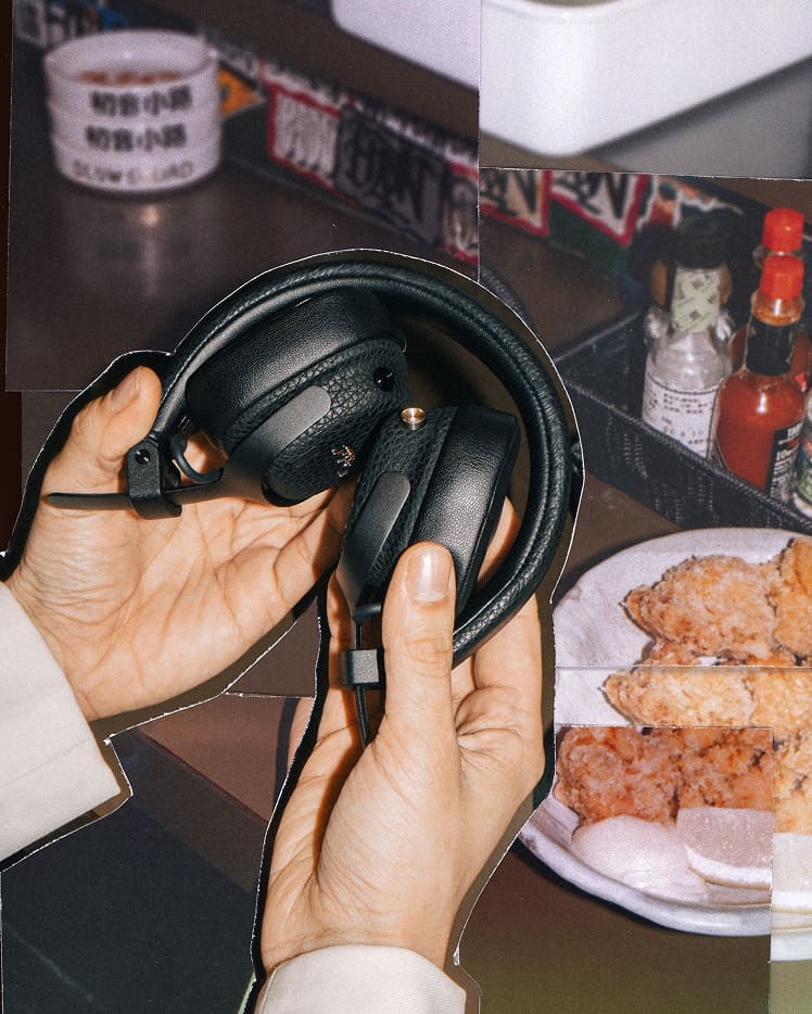A person holds Milton A.N.C. headphones in front of a table with fried food, condiments, and a cup in the background.