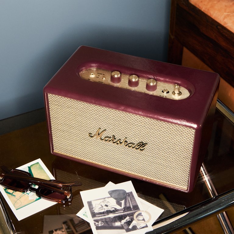Acton III Burgundy speaker with gold controls sits on a glass table beside sunglasses and black-and-white photos.