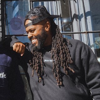 Troy D. Johnson, with long dreadlocks, a beard, and a black cap smiles while leaning on someone’s shoulder outside in sunlight.