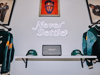 A shelf with two green "Never Settle" hats and a Marshall speaker below a neon "Never Settle" sign and framed portraits on a wall. Two green jackets hang on either side.