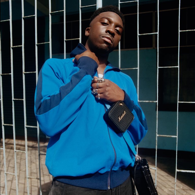 A man wearing a blue zip-up jacket and holding Marshall Willen II Black and Brass portable Bluetooth speaker.