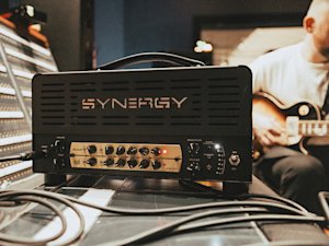 Un amplificateur Synergy avec le module de préamplification Marshall JCM800 Synergy est posé sur une table avec des câbles branchés ; une personne à l'arrière-plan joue d'une guitare électrique.