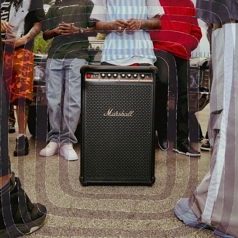 Five people stand behind a large Marshall Bromley 750 party speaker on the pavement, with part of a car visible in the background.