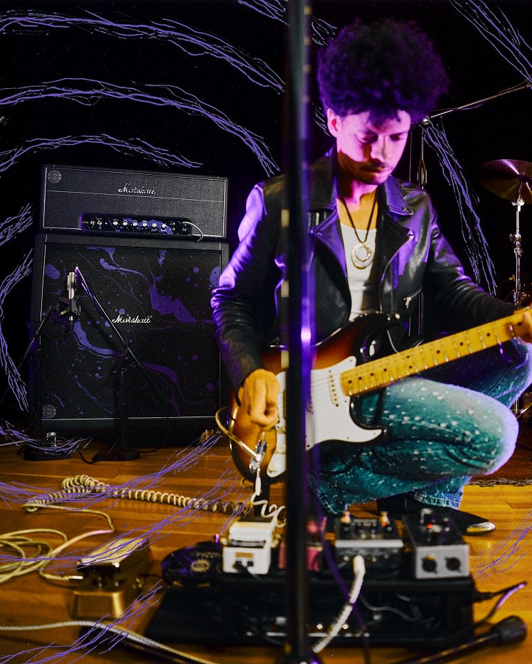 Un musicien est agenouillé sur scène, ajustant des pédales de guitare, tenant une guitare électrique, avec Marshall x Hendrix 60th Anniversary Collection en arrière-plan.