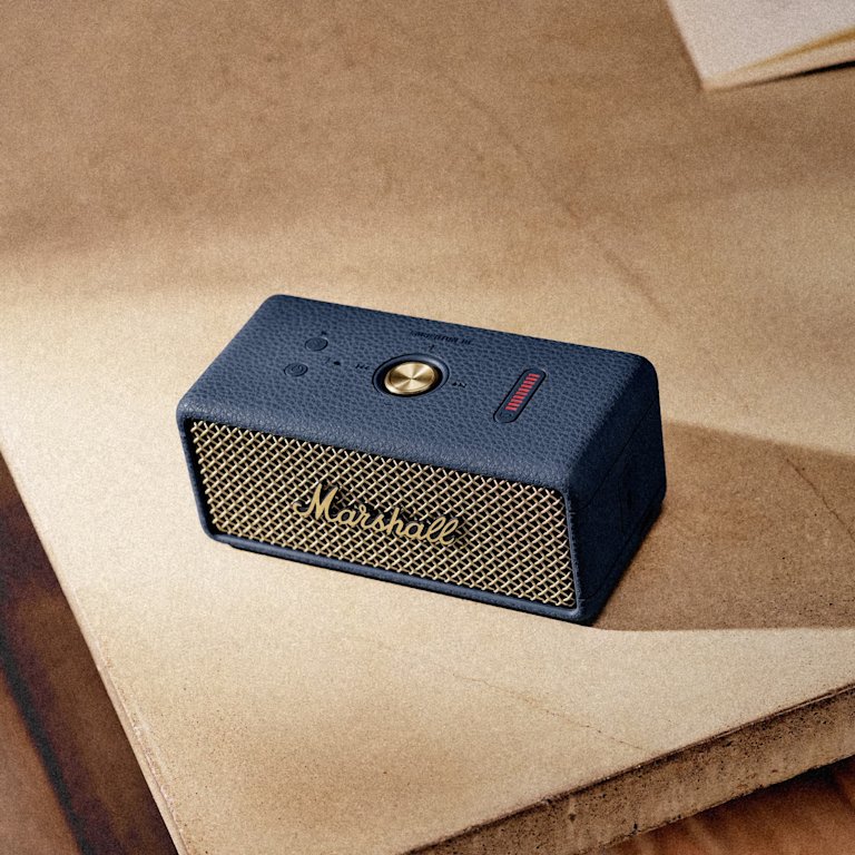 Marshall Emberton III Midnight Blue portable speaker sits on a wooden surface near a stack of papers, delivering powerful Bluetooth sound.