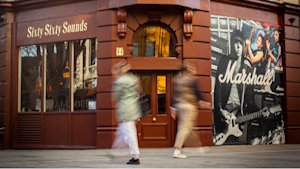 Two people walk past the Sixty Sixty Sounds storefront, which features a large Marshall advertisement with images of musicians and guitars.