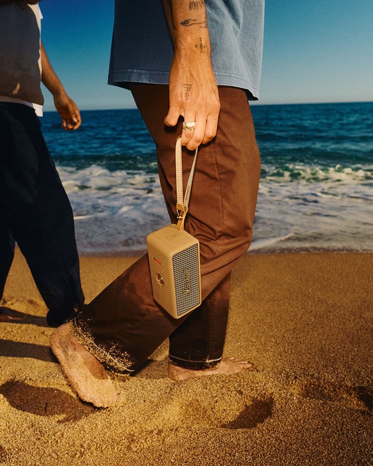 A person walks on the beach, holding an Emberton III Brown portable speaker by its strap.
