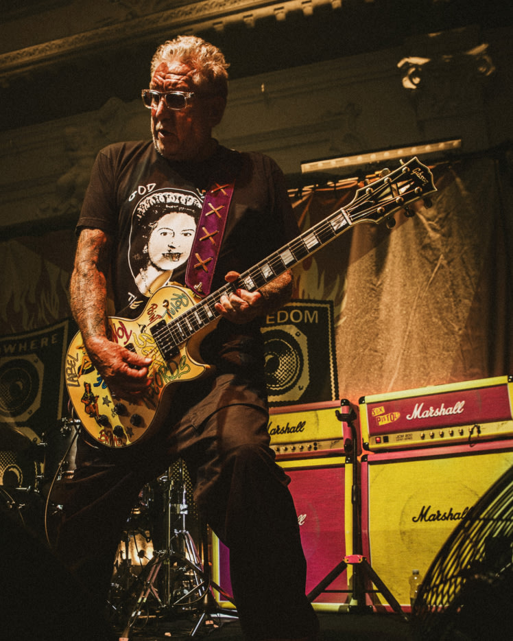 Eine Person spielt eine gelbe E-Gitarre mit den Verstärkern der Sex Pistols im Hintergrund