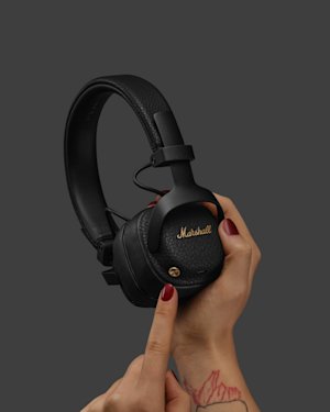 A hand with red nail polish holds black Marshall headphones against a gray background, with an index finger pointing to the earcup.