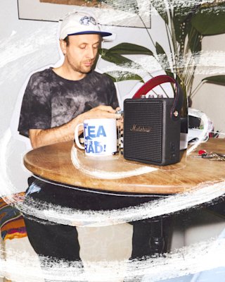 A man sits at a round table with a large mug, a Marshall Stockwell III Black and Brass speaker, and keys.
