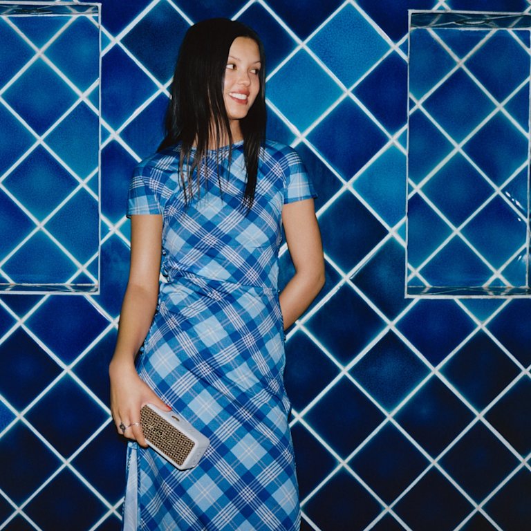 A woman in a blue plaid dress holding Marshall Emberton III Cream portable speaker.