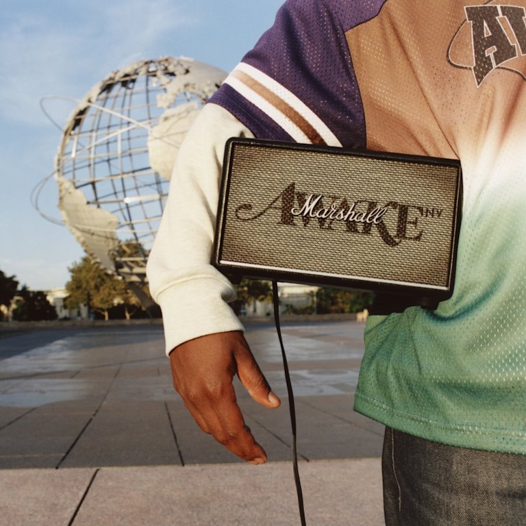 A person holding a Marshall Acton III Awake NY Edition home speaker under their arm stands near a globe sculpture, wearing a multicolored shirt.