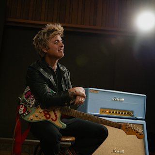 Billie Joe Armstrong in a black leather jacket sits on a stool holding an electric guitar, with a blue Marshall 1959 BJA amplifier in the background.