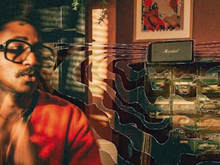 A man with orange sweater and glasses sits in a room with shelves and Marshall Acton IV Black speaker.