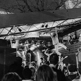 A black and white photo of a live outdoor concert, with one performer hanging from a roof beam above the stage while a crowd watches.